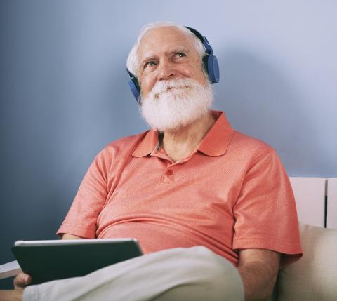 Een oudere man met witte baard heeft een hoofdtelefoon op en een tablet in zijn handen. Hij lijkt te genieten van wat hij hoort.