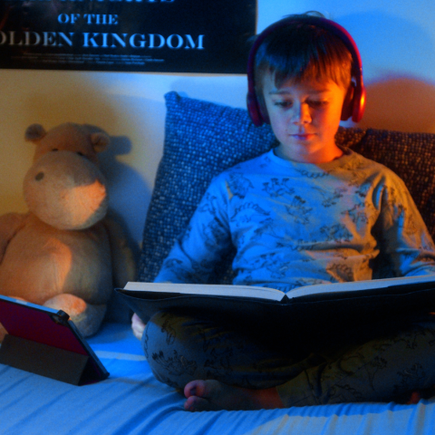 Jongen leest in bed in een boek met koptelefoon op en tablet erbij.