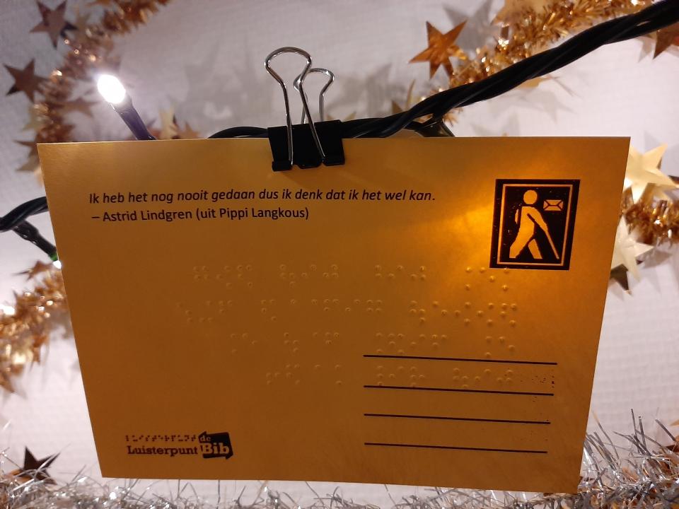 Een gele braillewenskaart van Luisterpunt met de tekst 'Ik heb het nog nooit gedaan dus ik denk dat ik het wel kan' (uit Pipi Langkous). Rond de wenskaart hangen kerstslingers en lichtjes. 