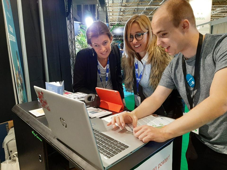 Een medewerker van Luisterpunt laat aan twee bezoekers van een beurs de mogelijkheden van Luisterpunt zien op een laptop 